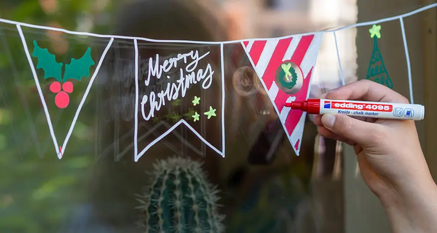 Eine junge Frau malt mit einem roten Kreidemarker von edding eine weihnachtliche Wimpelkette auf eine Fensterscheibe. Foto: Johanna Rundel