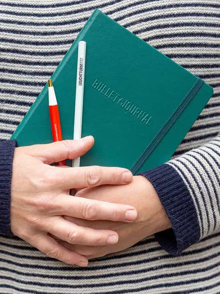 Eine Person in einem geringelten Wollpullover hält mit beiden Händen ein Bullet Journal von LEUCHTTURM1917, einen Drehgriffel und einen Bleistift vor ihren Bauch. Foto: Johanna Rundel