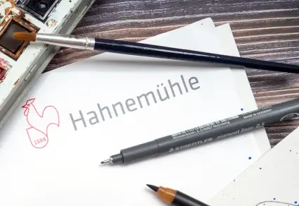 Das Hahnemühle-Logo auf weißem Grund zwischen verschiedenen Malutensilien.