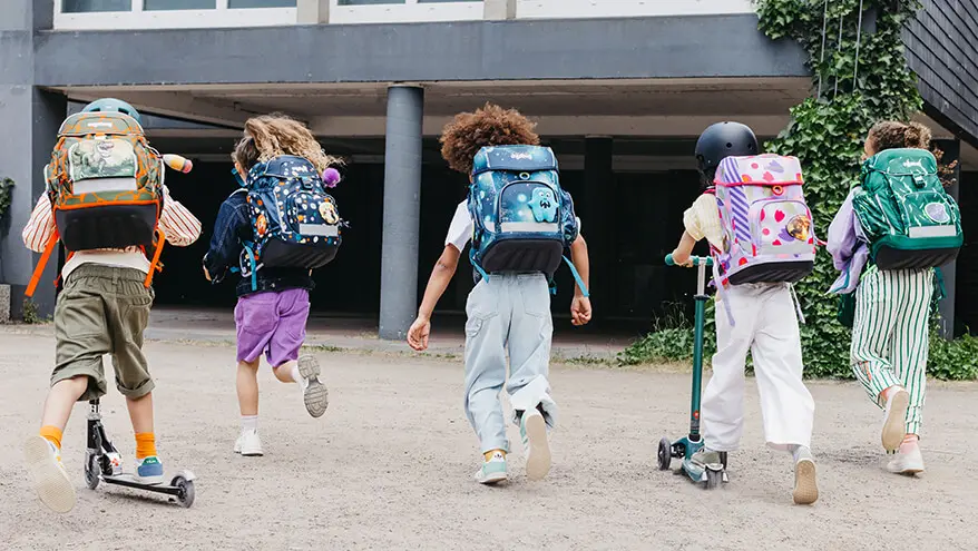 Fünf Grundschulkinder laufen mit ihren Schulranzen von ergobag auf dem Rücken freudig auf ein Schulgebäude zu. Foto: Johanna Rundel