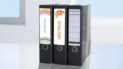 Drei schwarze Aktenordner mit verschieden gestalteten Ordnerrücken-Etiketten von Avery Zweckform stehen vor einem hellen Hintergrund.