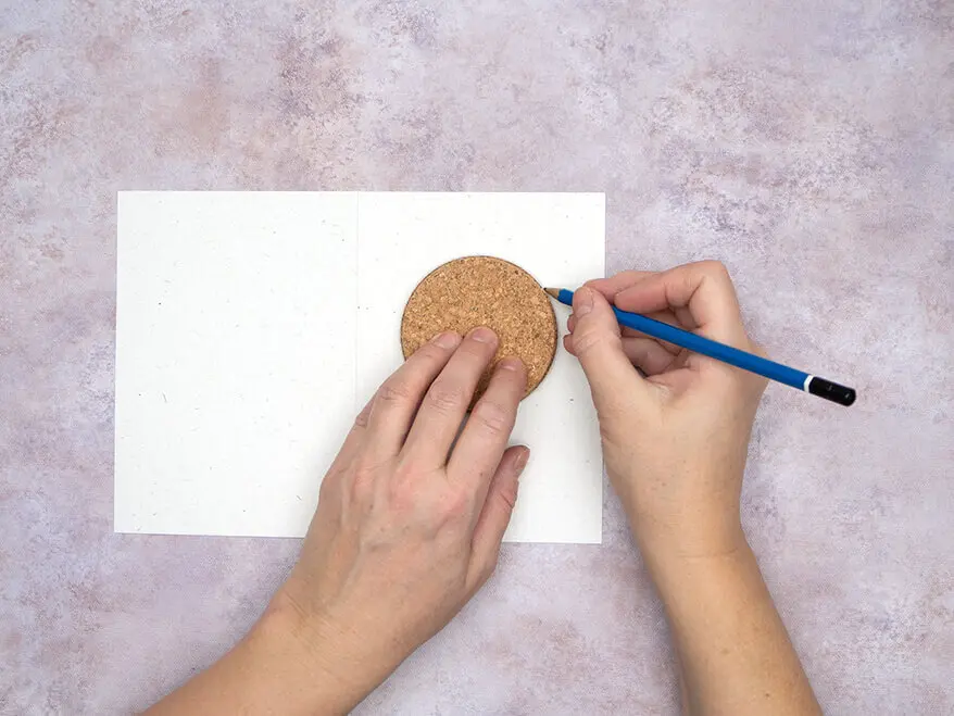 Jemand umfährt einen kleinen runden Korkuntersetzer mit einem Bleistift auf einer hellen Blanko-Klappkarte von Rössler. Foto: Johanna Rundel 