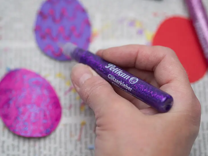 Jemand malt mit lilafarbigem Glitzerkleber von Pelikan Muster auf ausgeschnittene Eier aus buntem Tonpapier. Foto: Johanna Rundel