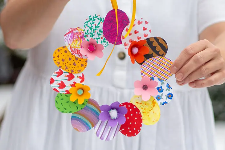 Eine Frau in einem weißen Kleid hält einen mit Materialien von Pelikan und herlitz gebastelten Osterkranz aus Papier in die Kamera. Foto: Johanna Rundel