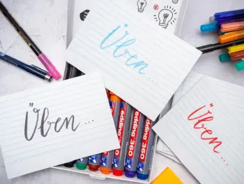 Drei Karteikarten auf denen „üben“ steht liegen auf diversen Stiften und Markern der Firma edding. Foto: Johanna Rundel