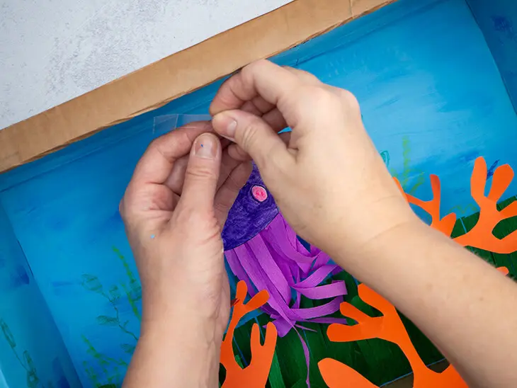 Jemand befestigt einen Fisch an einem Faden mit Klebefilm an die Decke eines DIY-Aquariums aus Karton. Foto: Johanna Rundel