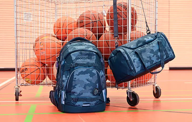 An einem Bällewagen mit Basketbällen in einer Sporthalle hängen eine Sporttasche und ein Schulrucksack von coocazoo.