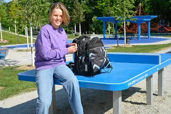 Ein Junge sitzt neben seinem Schulrucksack von coocazoo auf einer blauen Tischtennisplatte in einem Park.