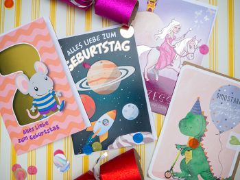 Vier verschiedene Geburtstagskarten für Kinder von ABC Grußkarten liegen nebeneinander auf einem gelb-weiß gestreiften Untergrund. Foto: Johanna Rundel