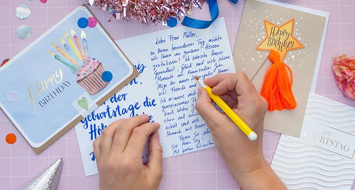 Jemand schreibt mit einem gelben Stift einen Text in eine Geburtstagskarte von ABC Grußkarten. Drumherum sind weitere Karten und Geburtstagsdeko verteilt. Foto: Johanna Rundel