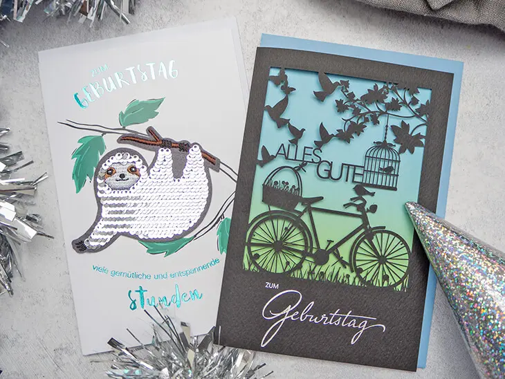 Zwei Geburtstagskarten mit Sonderausstattung (Scherenschnitt und Bügelbild) von ABC Grußkarten liegen auf einem grauen Untergrund. Foto: Johanna Rundel