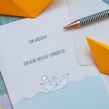 Eine Grußkarte mit Papierboot-Motiv und den Worten „zum Abschied alles Gute für deine weitere Lebensreise“ von Grafik Werkstatt. Daneben ein schwarz-weißer Kugelschreiber und gelbe Papierboote. Foto: Johanna Rundel