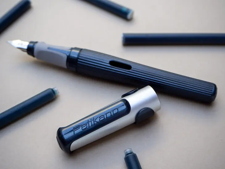 Ein schwarzer Pelikano-Füller von Pelikan mit feiner Reliefstruktur liegt zwischen verschiedenen Tintenpatronen auf einem grauen Untergrund. Foto: Johanna Rundel