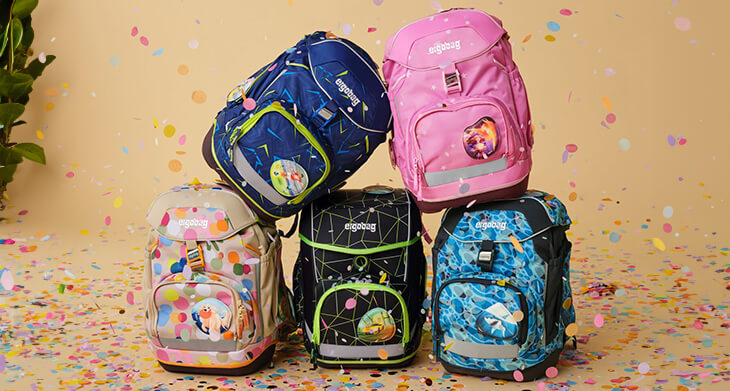 Fünf verschieden Schulranzen von ergobag stehen aufeinandergestapelt vor einem beigen Hintergrund. Sie sind von buntem Konfetti umgeben.