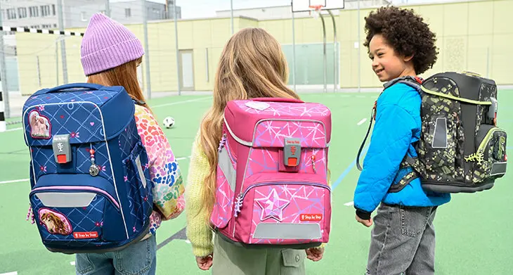 Drei Grundschulkinder stehen mit ihren Schulranzen von Step by Step auf dem Rücken auf einem Sportplatz einer Schule und unterhalten sich.