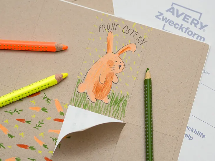 Ein Bogen Naturbraune Etiketten von Avery Zweckform, dessen Aufkleber mit Buntstiften mit Ostermotiven gestaltet wurde. Die Stifte liegen dabei. Foto: Johanna Rundel