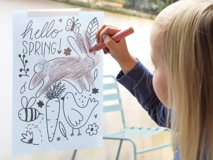 Ein kleines Mädchen mit blonden Haaren malt eine frühlingshafte Fensterbildvorlage mit einem STABILO woody-Buntstift von innen auf die Scheibe ab. Foto: Johanna Rundel