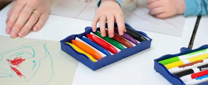 Eine Kinderhand greift in eine Box mit Wachsmalstiften, drumherum liegen bemalte Papiere