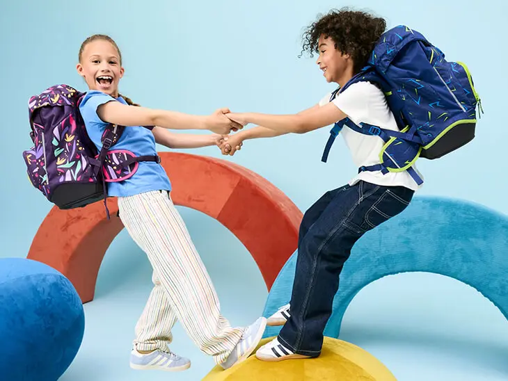 Ein Mädchen und ein Junge im Grundschulalter halten sich an den Händen und lehnen sich dabei zurück. Beide tragen einen Schulranzen von ergobag auf dem Rücken.