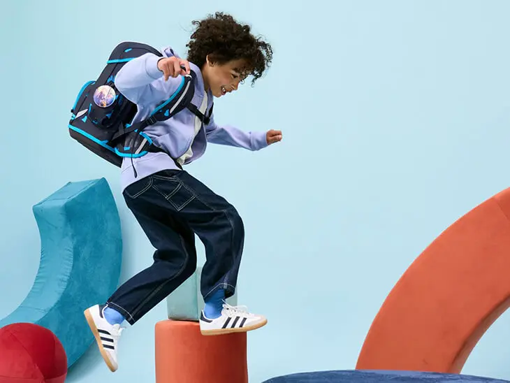 Ein Grundschüler springt mit seinem blauen Schulranzen von ergobag von einem Schaumstoffelement zum nächsten.)