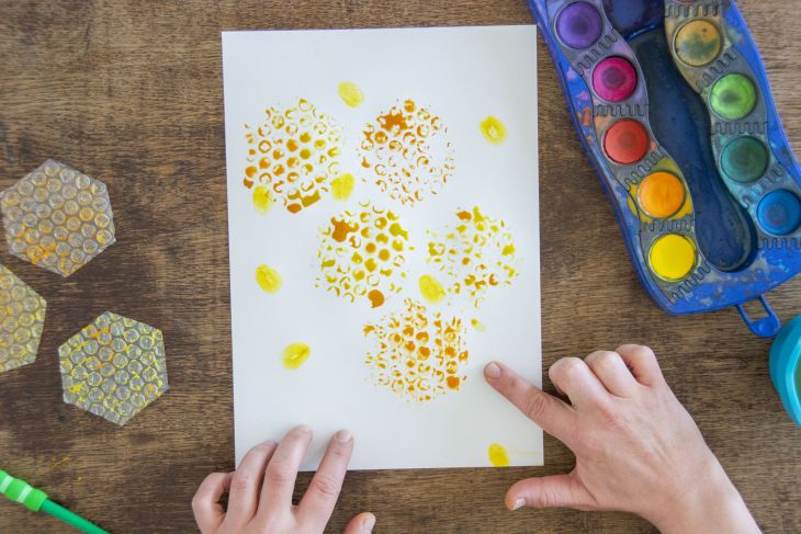 Jemand stempelt mit gelber Wassermalfarbe von Faber-Castell Fingerabdrücke auf ein Blatt Papier, auf dem Bienenwaben zu sehen sind
