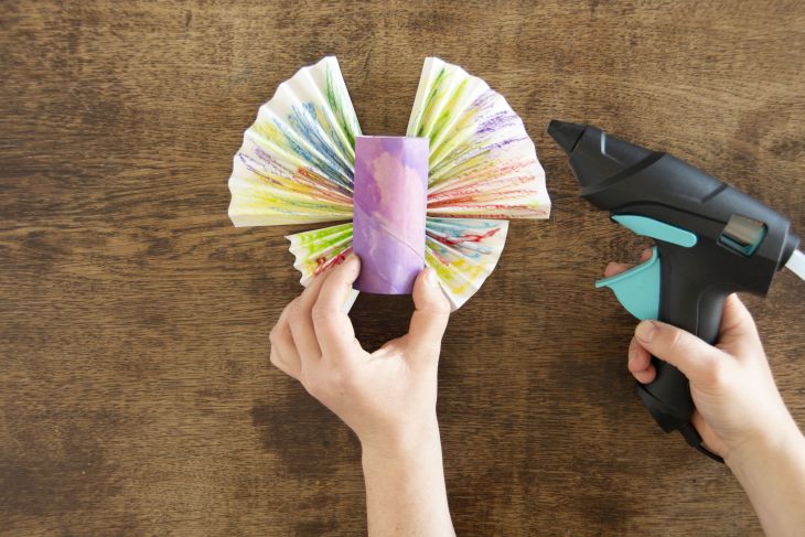 Jemand klebt selbstgefaltete Schmetterlingsflügel auf eine bemalte Klopapierrolle.