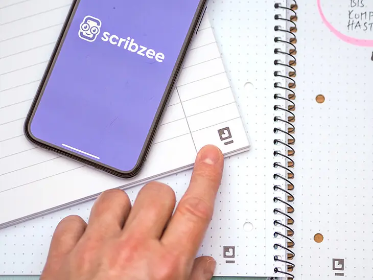 Jemand zeigt mit dem Finger in die Ecke eines Collegeblocks von Oxford auf ein Digitalisierungs-Icon. Die dazugehörige Scribzee-App ist im danebenliegenden Smartphone geöffnet. Foto: Johanna Rundel