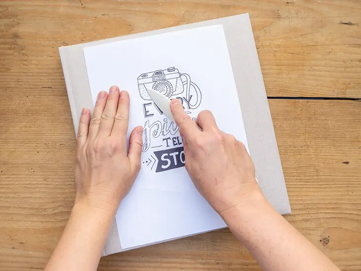 Jemand rubbelt mit einem Falzbein über eine Letteringvorlage auf einem Papier, um sie auf ein Fotoalbum-Cover zu übertragen. Foto: Johanna Rundel