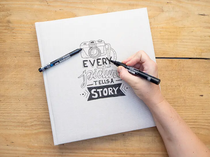 Jemand zeichnet eine Letteringvorlage „Every picture tells a story“ aus Bleistiftlinien auf einem Fotoalbum-Cover mit schwarzen Multimarkern nach. Foto: Johanna Rundel