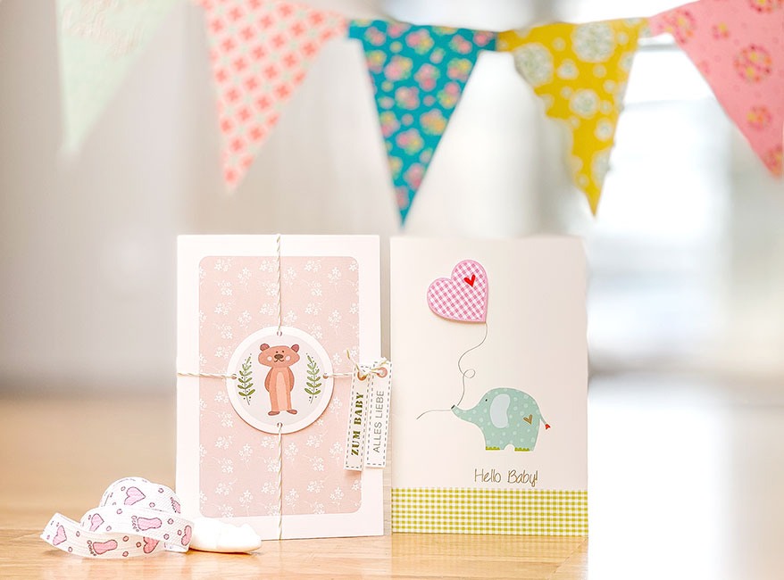 2 bunte Karten von Hanra zur Geburt stehen auf einem Tisch, darüber hängt eine bunte Girlande