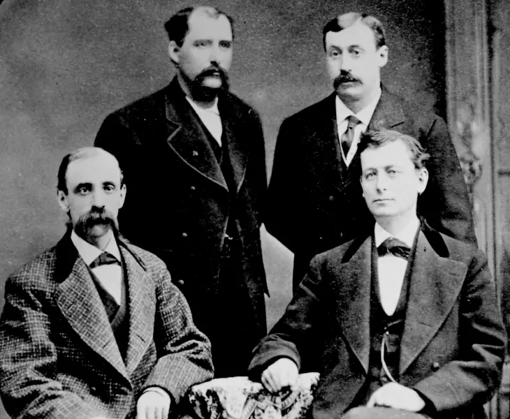 Schwarz weiß Foto von den vier Gründungsmitgliedern von Kimberly-Clark. John A. Kimberly und Frank Shattuck sitzen, während Charles Clark und Havilah Babcock hinter den Stühlen stehen. Alle tragen Krawatten und Anzüge und machen einen formellen Eindruck. Die Bildkennzeichnung lautet „Courtesy of Kimberly-Clark's Legacy“.