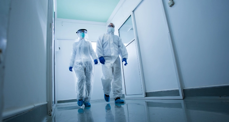 Zwei Personen in vollständiger Schutzausrüstung, inklusive Schutzanzügen und Masken, gehen durch einen sterilen Flur.