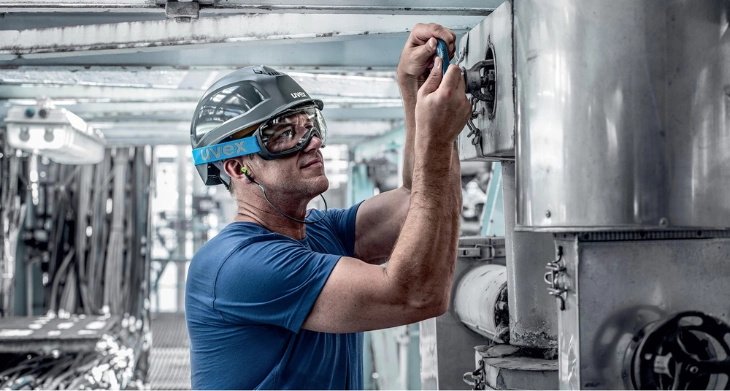 Arbeiter mit Helm, Schutzbrille und Gehörschutz repariert technische Anlage in einer Industriehalle.