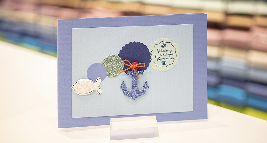 Eine selbstgebastelte Kommunionskarte steht in einem Aufsteller auf einem Tisch. Im Hintergrund ist verschwommen eine Papierwand mit vielen bunten Papieren zu sehen.