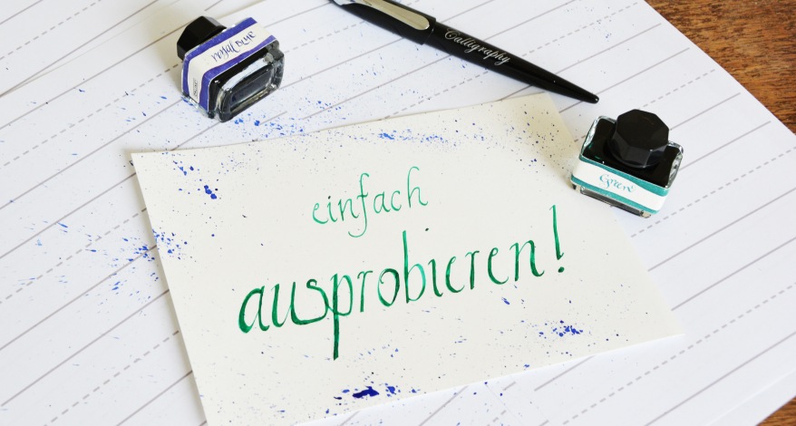 Auf einem linierten Papier liegt eine Karte, auf der mit grüner Tinte in Kalligarfie-Schrift "einfach ausprobieren!" steht. Daneben stehen zwei kleine Tintenfäasser und es liegt ein Kalligrafie-Stift daneben.