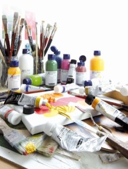 Auf einem Tisch liegen ganz viele Tuben Farbe der Marke Schmincke auf einer bemalten Leinwand. Es stehen Farben in kleinen Flaschen auf dem Tisch und Pinsel stehen in Gläsern daneben.