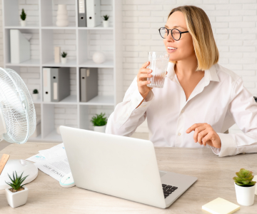 Frau sitzt an Schreibtisch vor ihrem Laptop und einem Ventilator mit einem Glas Wasser in der Hand