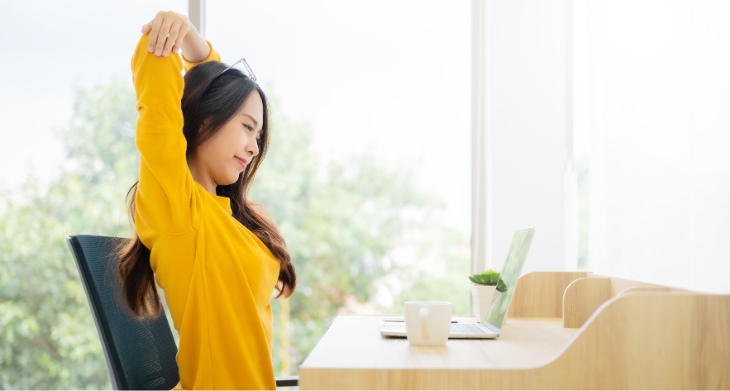 Das Bild zeigt eine Frau in einem gelben Pullover, die an ihrem Schreibtisch sitzt und Dehnübungen macht. Sie streckt ihre Arme nach oben, während sie lächelt und eine kurze Pause einlegt. Auf dem Tisch stehen ein Laptop, eine Tasse und eine kleine Pflanze. 