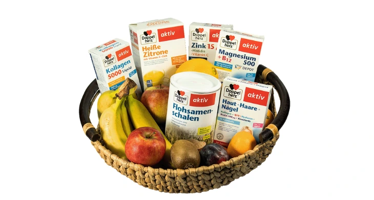 Das Bild zeigt einen geflochtenen Korb, der mit frischem Obst wie Bananen, Äpfeln, Pflaumen und einer Kiwi gefüllt ist. Zusätzlich enthält der Korb verschiedene Nahrungsergänzungsmittel der Marke Doppelherz, darunter Produkte wie Flohsamenschalen, Magnesium + B12, Zink, Kollagen und Haut-Haare-Nägel Tabletten für eine gezielte Nährstoffbersorgung..