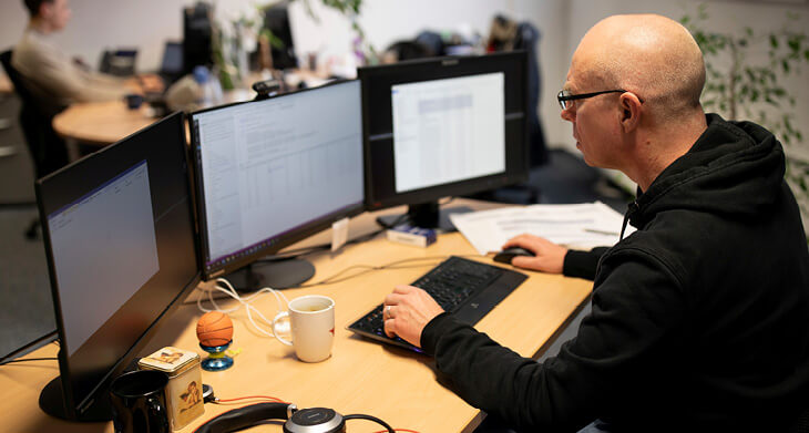 Ein Mann mit Glatze, Brille und schwarzem Kapuzenpullover sitzt in einem Büro. Man blickt seitlich auf den Mitarbeiter und seinen Schreibtisch, auf dem drei Monitore und eine Tastatur stehen. Außerdem stehen eine Tasse und mehrere kleine Gegenstände auf dem Schreibtisch. 