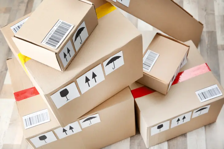 Die Abbildung zeigt Versandkartons in verschiedenen Größen, teilweise mit rotem und gelben Packband verschlossen und mit Hinweisetiketten “Bruchgefahr“, „Ware aufrecht stellen“ und „ Feuchtigkeitsempfindlich