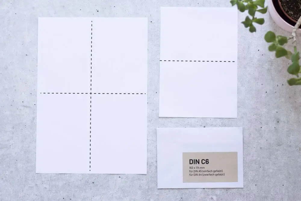 Auf grauem Grund liegt ein DIN A4-Papier mit zwei gestrichelten Markierungen, die das Papier in Viertel teilt und ein DIN A5-Papier mit einer gestrichelten Markierung in der Mitte, dazu ein passender Briefumschlag C6. Auf dem Briefumschlag ist ein Aufkleber aufgebracht mit folgenden Infos: DIN C6, 162x114 Millimeter, für DIN A5 (einfach gefalzt), für DIN A4 (zweifach gefalzt). Rechts oben ragt der Teil einer Pflanze ins Bild.