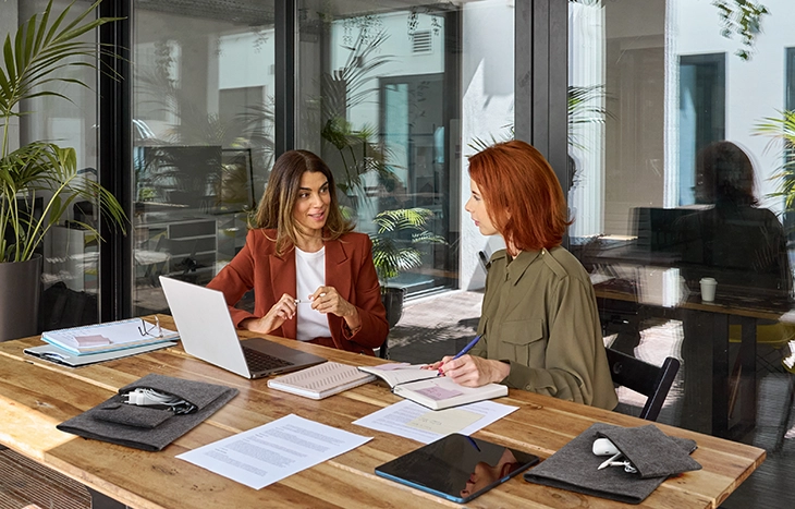 Zwei Frauen im Gespräch an modern gestaltetem Arbeitsplatz mit Laptop.