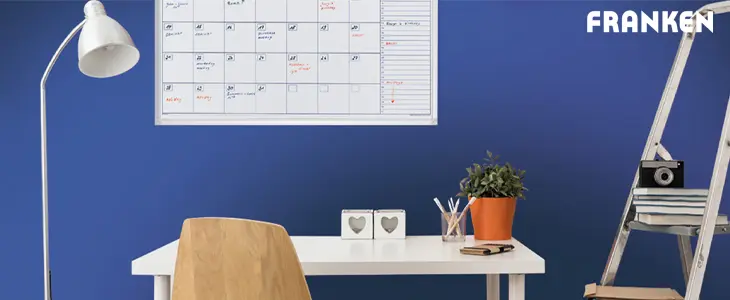Das Bild zeigt einen minimalistischen Arbeitsplatz mit einem weißen Schreibtisch, einer Lampe und einer Pflanze. An der Wand hängt eine Plantafel, die eine klare Übersicht über Termine und Aufgaben bietet. Neben dem Tisch steht eine Leiter mit gestapelten Büchern und Unterlagen.