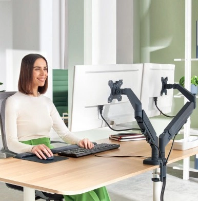 Das Bild zeigt eine Frau, die an einem Schreibtisch mit zwei Monitorhaltern arbeitet, die auf Augenhöhe eingestellt sind. Sie sitzt in ergonomischer Haltung und nutzt eine Tastatur und Maus.