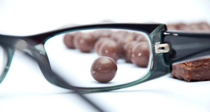 Das Bild zeigt eine Lesebrille, durch deren Glas mehrere schokoladenüberzogene Kugeln scharf zu sehen sind, während der Rest des Bildes unscharf bleibt. 