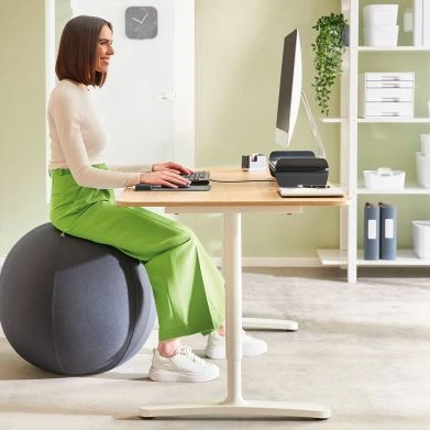 Das Bild zeigt eine Frau, die an einem Schreibtisch arbeitet und dabei auf einem Sitzball sitzt. Sie hat eine aufrechte Haltung, während sie auf den Monitor schaut und tippt. 