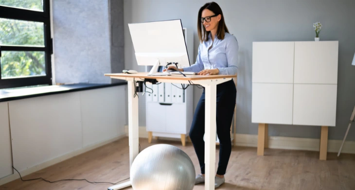 Das Bild zeigt eine Frau, die an einem höhenverstellbaren Stehschreibtisch steht und dabei auf einem Monitor schaut. Unter dem Tisch liegt ein silberner Gymnastikball für mehr Bewegung und ergonomische Alternativen im Büro. Die Szene betont die Bedeutung von Ergonomie, Stehlösungen und gesunder Haltung am Arbeitsplatz.