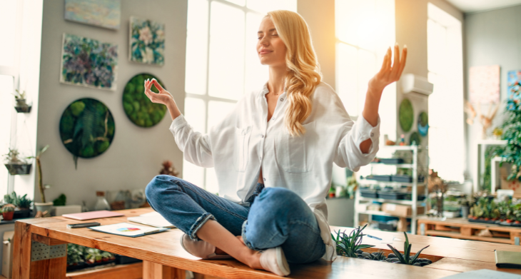 Das Bild zeigt eine lächelnde junge Frau mit langen blonden Haaren, die entspannt im Schneidersitz auf einem Schreibtisch sitzt und meditativ die Hände in einer Atemübung-Pose hält. Um sie herum stehen Pflanzen, Bücher und Deko-Elemente, was eine ruhige Atmosphäre schafft. 