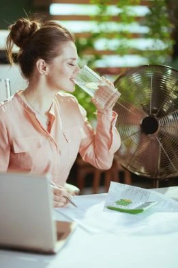Das Bild zeigt eine Frau in einem Büro, die ein Glas Wasser trinkt. Sie sitzt an einem Schreibtisch mit einem Laptop und Dokumenten, während ein Ventilator im Hintergrund steht. Die Szene betont die Wichtigkeit von ausreichend Trinken für die Gesundheit am Arbeitsplatz. 